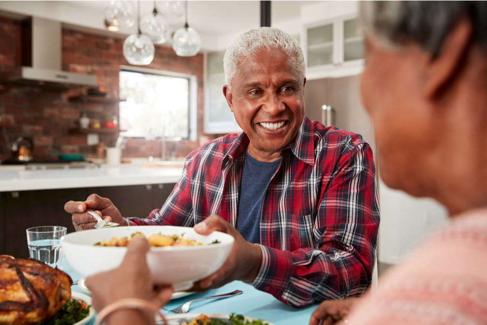 Two seniors pass a plate of food at the dinner table.